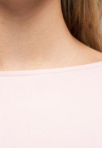 Κοντινό πλάνο του λαιμού και του άνω μέρους του στήθους ενός ατόμου που φοράει ροζ ανοιχτό, μπλουζάκι με στρογγυλή λαιμόκοψη, με ξανθά μαλλιά ορατά στο πλάι.