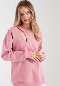 Różowa bluza z unikalnym wycięciem i miękką fakturą. Długie rękawy z prążkowanymi mankietami i luźny krój. Prosty, nowoczesny styl.