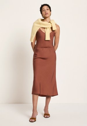 Frau trägt ein ärmelloses, rostfarbenes Midikleid, beiger Pullover über den Schultern drapiert, braune Sandalen mit offenen Zehen, steht vor einem schlichten Hintergrund.