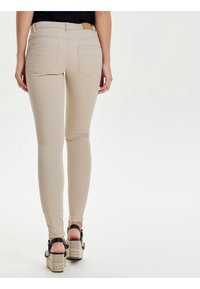 Mujer vestida con jeans ajustados beige y sandalias de cuña negras, de pie con la espalda hacia el espectador sobre un fondo blanco.