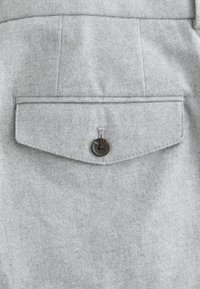Poche à rabat boutonnée sur tissu texturé gris clair, montrant des coutures détaillées et un bouton foncé à quatre trous.