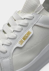 Λευκό αθλητικό παπούτσι με υφασμάτινη υφή, χοντρές λευκές κορδόνια και την επιγραφή "LOVE MOSCHINO" σε χρυσά γράμματα στην πλαϊνή επιφάνεια.