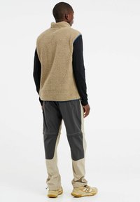 Beige fleece vest met hoge kraag over een zwart langemouwshirt; gecombineerd met grijze en beige broek. Textuur, casual stijl.