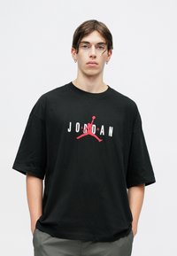 Crna majica s kratkim rukavima koja na prednjoj strani ima crveno-bijeli Jordan logo. Širok kroj, okrugli izrez. Minimalistički dizajn.