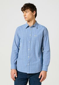 Camisa de manga larga a cuadros azules, hecha de una tela ligera, con cuello abotonado y un bolsillo de parche en el lado izquierdo del pecho.