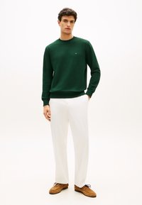 Πράσινο πουλόβερ με μακριά μανίκια και στρογγυλή λαιμόκοψη, μαλακή υφή και ριμπ μανσέτες, συνδυασμένο με λευκό παντελόνι και καφέ παπούτσια από σουέτ.