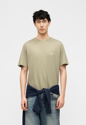 Jeune homme aux cheveux noirs bouclés, portant un t-shirt beige avec le texte "ORIGINALS" et un pull bleu marine noué autour de la taille.