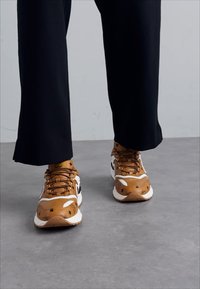 Baskets marron et blanches avec des accents noirs, présentant une partie supérieure texturée, un design à lacets et une semelle blanche épaisse, associées à des pantalons noirs.