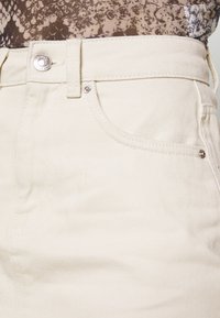 Närbild av beige denimbyxor i midjan med metallknapp, framficka och bältesöglor, som bärs med topp i ormskinnsmönster.