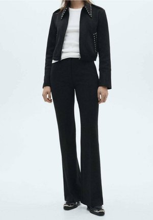 Personne portant une veste courte noire à clous, un haut blanc, un pantalon noir évasé taille haute et des chaussures noires, debout devant un fond uni.