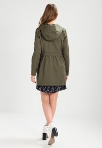 Olivgrön huva jacka med insnörad midja, långa ärmar och en bakre fåll som slutar vid mitten av låret, matchad med en mörkblommig kjol.