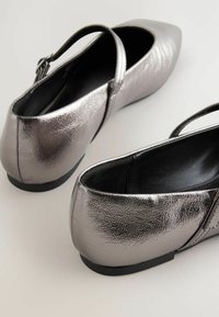 Metallisch zilveren ballerina's met een textuurafwerking, puntige neus en verstelbare band. Zwarte binnenkant en een lage zool.