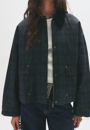 Kvinde iført en mørkegrøn og marineblå ternet jakke med klaplommer, over en hvid skjorte og en ternet indertrøje, kombineret med mørkeblå jeans.