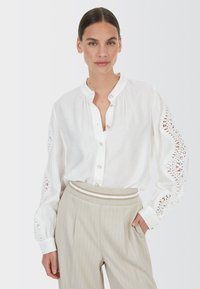 Weißes Hemd mit langen Ärmeln, das Spitze an den Schultern hat. Kombiniert mit hellbeigen, gestreiften Hosen.