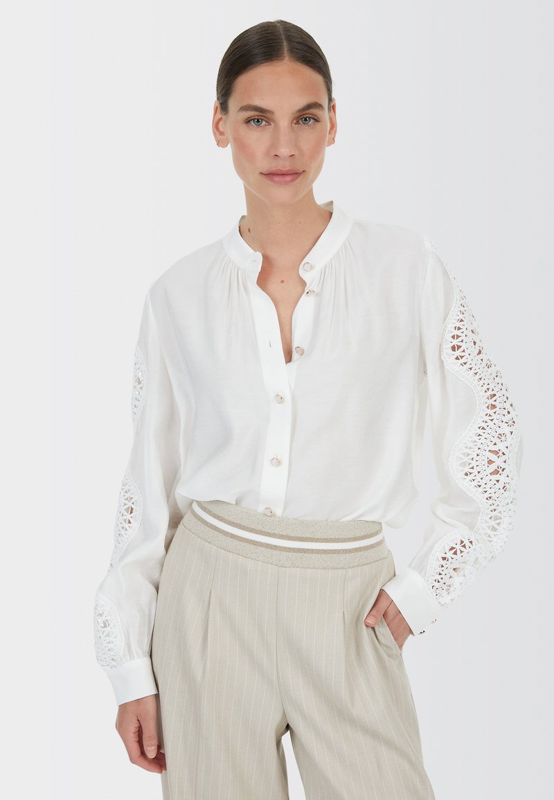 Weißes Hemd mit langen Ärmeln, das Spitze an den Schultern hat. Kombiniert mit hellbeigen, gestreiften Hosen.