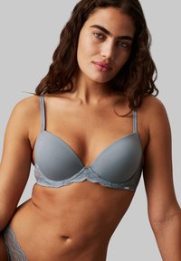 Gray padded bra with lace trim and adjustable straps. Smooth texture with scalloped edges and subtle sheen. Model posed in a neutral setting.