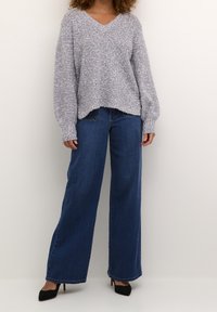 Pull gris en maille avec col en V, manches longues et amples, associé à un jean en denim bleu taille haute à jambes larges et des talons noirs pointus.