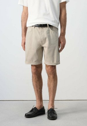 Mężczyzna ubrany w beżowe szorty, białą koszulkę, czarny pasek i czarne buty casual stojący na jednolitym tle.