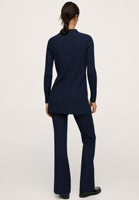 Žena s tmavými vlasy v culíku, oblečená v námořnickém dlouhém rukávu s ribováním a odpovídajících kalhotách, stojí zády k nám na jednobarevném pozadí.