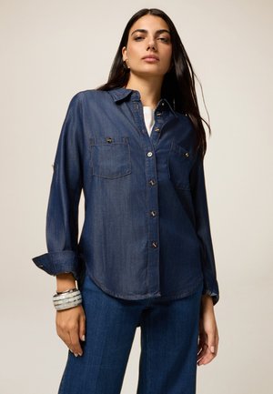 Dark denim shirt with a button-up design, two chest pockets, and rolled sleeves. Features a curved hem and silver-tone buttons.