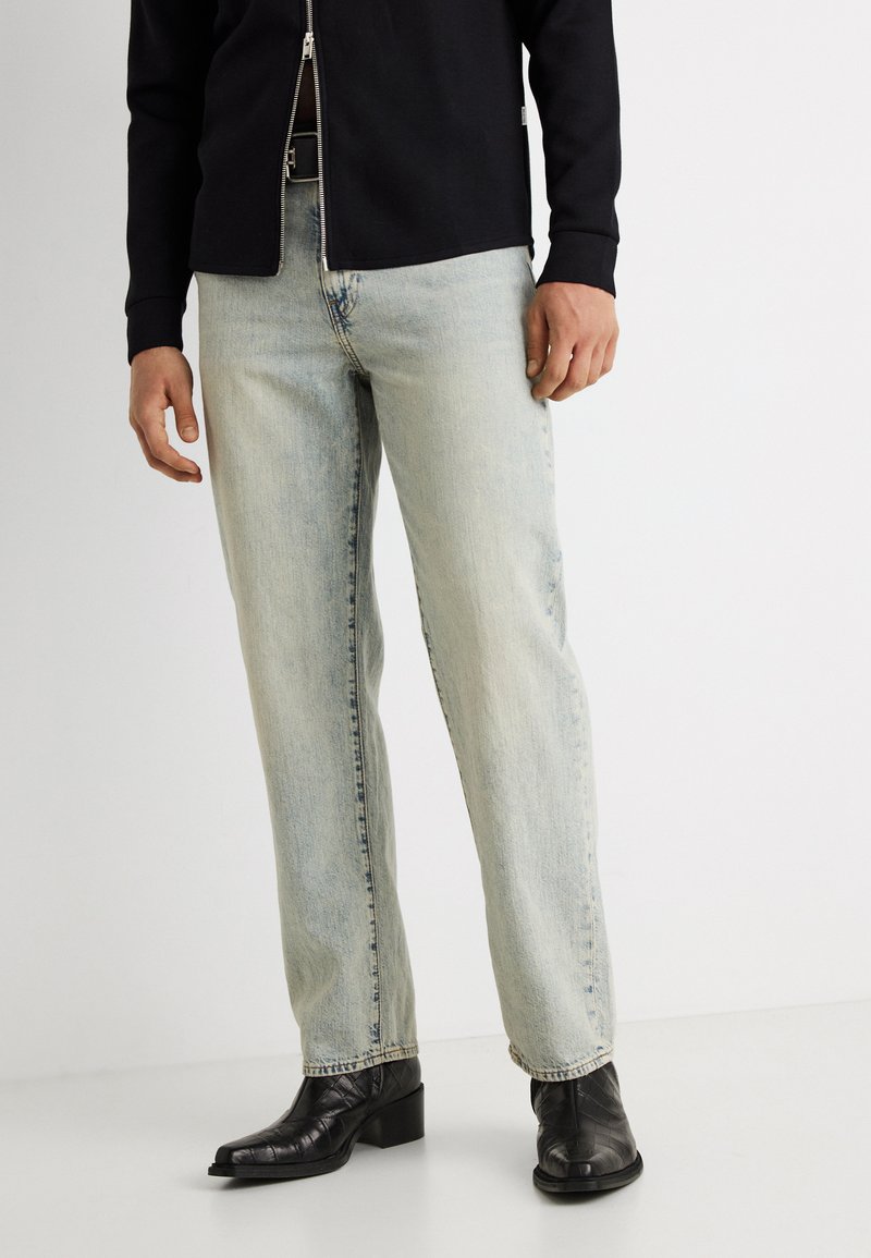 Άνδρας που φοράει φθαρμένα τζιν με ίσια μπατζάκια σε ανοιχτό γαλάζιο χρώμα, μαύρο τζάκετ με φερμουάρ και μαύρα δερμάτινα μποτάκια αστραγάλου.