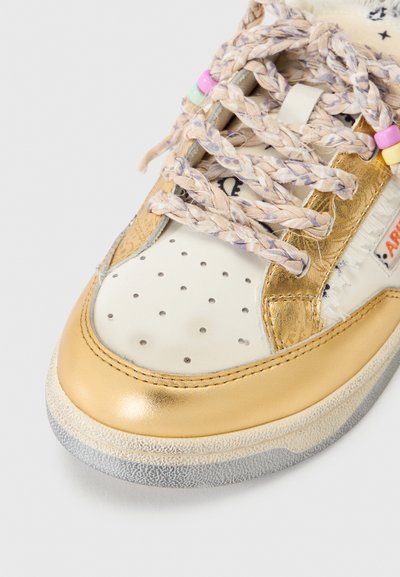 Λευκό και χρυσό αθλητικό παπούτσι με διάτρητη μύτη, πλεξούδες πολύχρωμα κορδόνια και μικρές χάντρες ως διακοσμητικά σε λευκό φόντο.