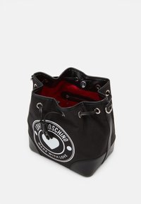 Svart tyg-hinkväska med röd inredning, med en cirkulär vit logotyp och dragsko stängning med silver-oöglor och svarta läderdetaljer.