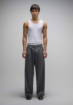 Jeune homme aux cheveux courts et foncés portant un débardeur blanc, un pantalon gris ample et des chaussures noires, debout devant un fond gris uni.