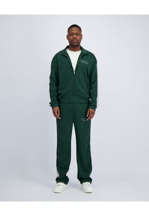 Man staat en draagt een donkergroen trainingsjack en broek met witte strepen en witte sneakers tegen een effen grijze achtergrond.