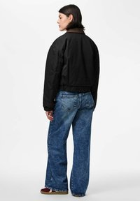 Kobieta z krótkimi ciemnymi włosami, ubrana w czarną kurtkę z brązowym kołnierzem, luźne niebieskie dżinsy i bordowe sneakersy, stojąca w profilu na białym tle.