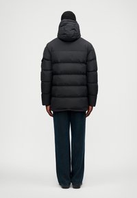 Άτομο που στέκεται με την πλάτη γυρισμένη, φορώντας μαύρο γεμιστό μπουφάν με κουκούλα και σκούρους φαρδείς παντελόνες, σε απλό φόντο.