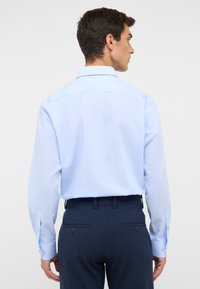 Camicia azzurra a maniche lunghe con colletto a punta, texture liscia e polsini abbottonati, indossata con pantaloni scuri che mostrano tasche posteriori e bottoni.