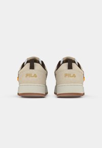 Beige semskinn sneakers med lav profil, som har en hvit såle, kontrasterende mørkebrune detaljer og en oransje etikett. Fila-logo i gull.