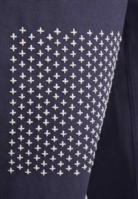 Tkanina mornarsko modre barve vsebuje vzorec belega križnega vezenja, ki dodaja teksturo in podrobnosti. Material deluje trpežno z urejeno zasnovo.