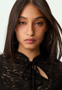 Blusa nera in pizzo con dettaglio alla scollatura, caratterizzata da un'apertura a forma di chiave. La modella ha i capelli lunghi e scuri e indossa grandi orecchini a cerchio dorati.