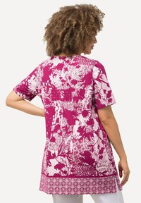 Kurzarmlige Tunika in Magenta mit weißen abstrakten Mustern, mit abgerundetem Saum und dekorativer Borte am unteren Rand.