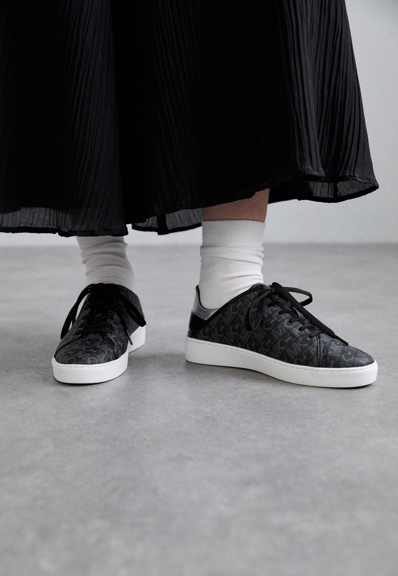 Personne portant des chaussettes blanches et des baskets noires à motifs avec des semelles blanches, debout sur un sol gris sous une jupe plissée noire.