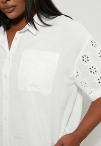 Camicia bianca di lino a maniche corte, con ricami in pizzo dettagliati su maniche e tasca, dotata di colletto classico e chiusura frontale con bottoni.