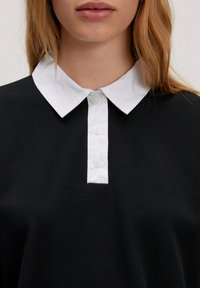 Zwarte polo met een witte kraag, met vijf knopen aan de voorkant en een gestructureerde stof.