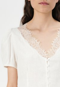 Hvit bluse med V-hals med blondekant, korte puffærmer og knappelukking. Materialet ser lett ut med en teksturert finish.