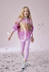 Glitrende lyserød sweater med pailletter og guldaccenter, parret med skinnende lyserøde leggings og hvide sneakers; sølvkonfetti spredt ud over gulvet.