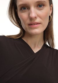 Brązowa, miękka bluzka z rozciągliwego materiału z opaską. Posiada głęboki dekolt w kształcie V i gładką teksturę. Ubrane są złote kolczyki w kształcie kół.