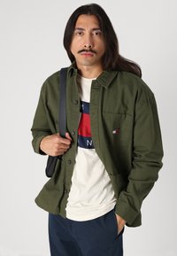 Chemise olive à manches longues avec une poche poitrine, portée sur un T-shirt léger à rayures horizontales bleu, rouge et blanc.