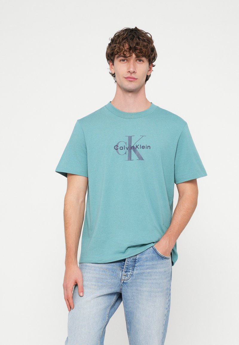 Turkio spalvos medvilniniai marškinėliai su tamsiai violetiniu atspaustu „Calvin Klein“ logotipu. Klasikinio kirpimo apvali kaklo iškirptė, trumpomis rankovėmis, laisvo stiliaus.