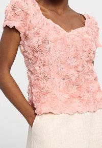 Haut floral rose en tissu texturé avec des appliqués de rose superposés. Manches courtes et décolleté en V. Associé à un bas de couleur claire.