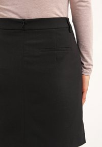 Falda lápiz negra con una textura suave, que presenta un cierre de cremallera escondido y un bolsillo trasero. Ajuste entallado y diseño hasta la rodilla.