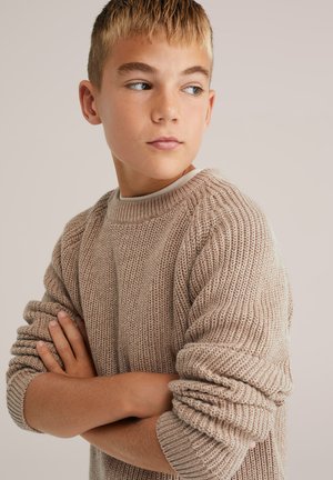 Bežs adīts džemperis ar ribotu tekstūru, apaļu kakla izgriezumu un sarullētām piedurknēm, ko valkā jauna modele ar rokām krustā.