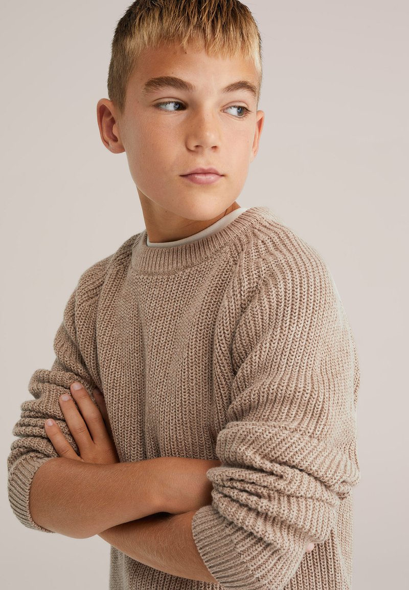 Bež pleten pulover z ribasto teksturo, okroglo ovratnikom in zvitimi manšetami, nosi mlad model z crossed arms.