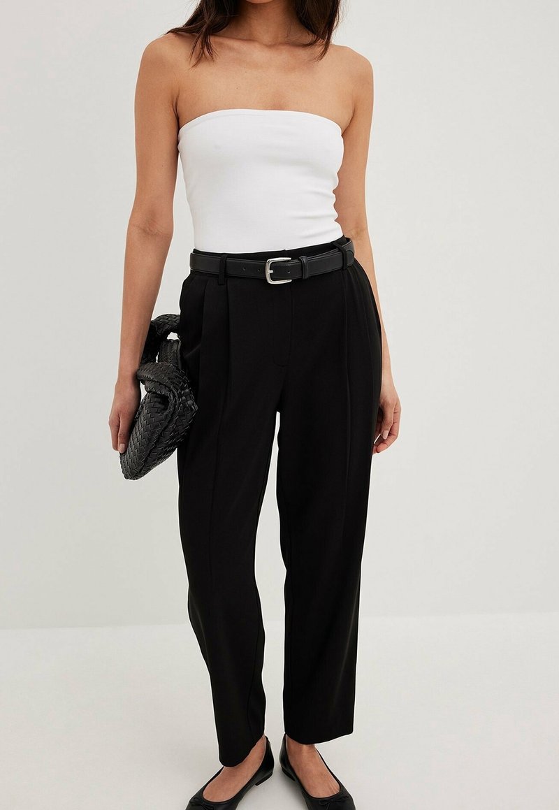 Femme portant un haut bustier blanc, un pantalon noir taille haute avec ceinture, tenant une pochette noire tissée et des chaussures plates noires sur un fond neutre.