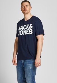 Navy blue cotton t-shirt featuring large white text "JACK&JONES," with a relaxed fit and short sleeves. Worn with distressed blue jeans.